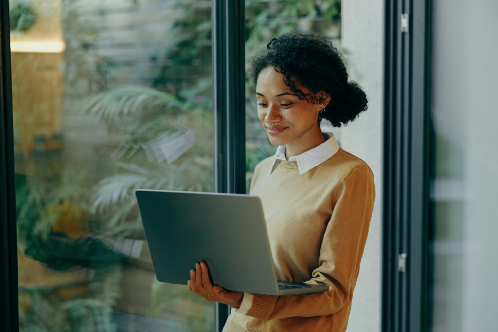 Female freelancer working laptop remotely while standing near window at home. Distance work concept